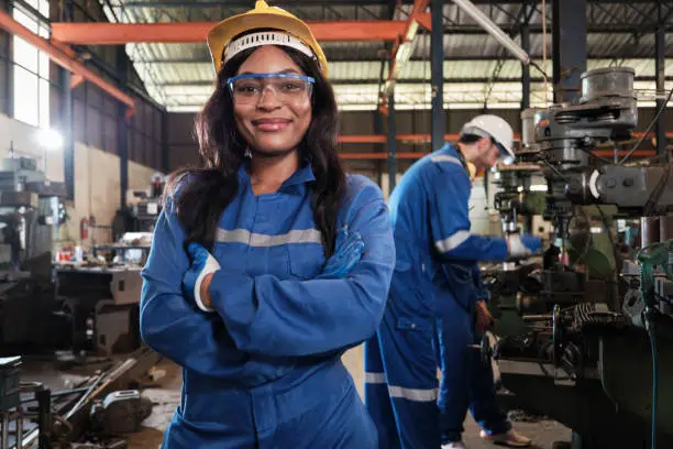 Portrait of professional Black female worker in protective safety uniform and helmet looking at camera, arms crossed and smiling, with engineers team behind her in a metalwork manufacturing factory.