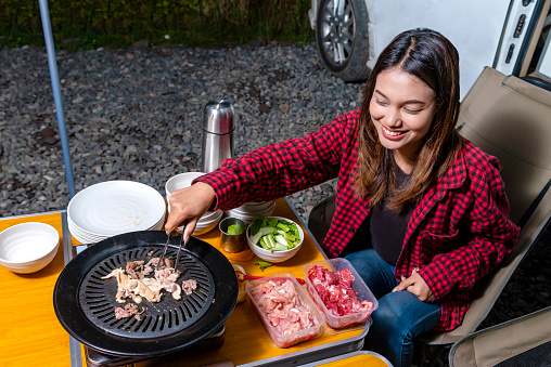 Asian woman sitting while grilling barbeque enjoying camp activity in nature