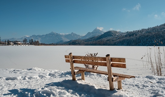 A wooden bench by the frozen Weissensee lake before the mountains on a sunny winter day