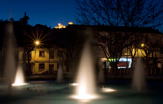 Verín town square, illuminated ornamental fountain, Verín town, Ourense province, Galicia, Spain. Monterrei defensive towers illuminated on the background mountain.
