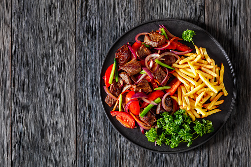 Lomo saltado, Peruvian dish of stir-fried slices of beef sirloin, onions, yellow Peruvian chilis, and tomatoes served with French fries on black plate on dark wood table, flat lay, free space