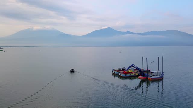 Drone shot of huge lake with fisherman boat crossing on the side of dredger boat. Seen sunrise sky and mountain on background - Rawa Pening Lake, Central Java, Indonesia