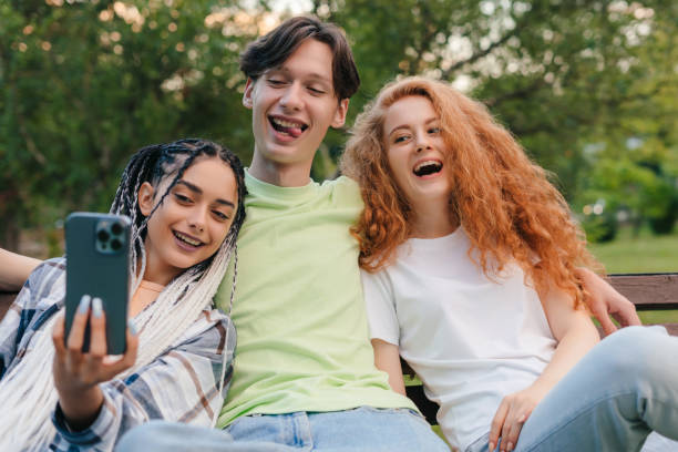 istock Three caucasian smiling friends at the park sitting on a bench and taking selfies using a smart phone. Summer vacation fun. Having fun. 1449298416