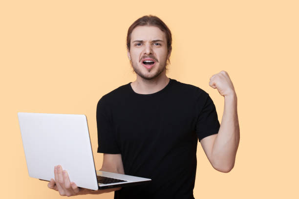 istock Portrait of a smiling young handsome man looking at camera while holding laptop, doing a winning closed fist gesture isolated over beige background. Positive person. Technology concept. 1449301466