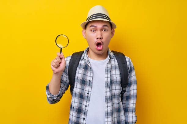 istock traveler fun explorer amazed tourist Asian man in casual clothes hat with backpack using magnifier loupe isolated on yellow background. Air flight journey concept 1449302223