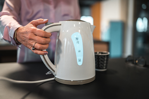 Close up on hands of a Caucasian female employee making herself a cup of tea at work. Boling a water in an electric kettle.