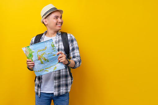 Smiling young traveler tourist Asian man wearing casual clothes and hat with a backpack holding city map, looking aside isolated on yellow background. Air flight journey concept