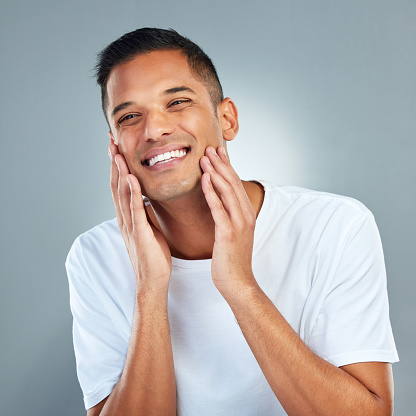 Man, smile and face with skincare, beauty and dermatology in studio for cleaning, hygiene and healthy skin. Model, happy and hands on cheeks for health, wellness and cosmetics against grey background