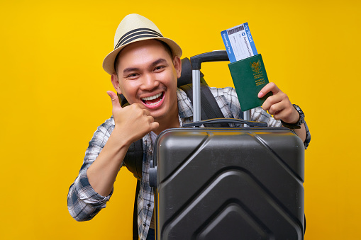 Smiling young Traveler tourist Asian man in casual clothes and backpack with a hat suitcase holding passport, boarding pass ticket, showing thumbs up on yellow background. Air flight travel concept