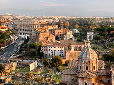 High angle view of Rome's historic downtown with the Coliseum surrounded by the old ruins of the Roman Forum, all bathed by warm sunlight at sunset. Dome, churches, trees and tourists walking  on the nearby streets.