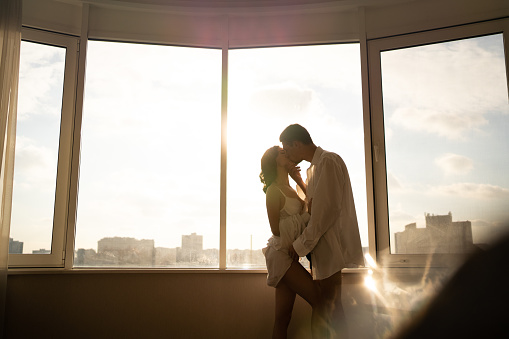 Woman and man hugging and kissing by the panoramic window at hotel. Festive morning, engagement party, happy couple, Valentines day celebration, romance and love concept.