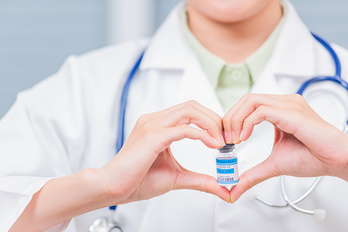 Closeup woman doctor smile forming a heart with her hands mark heart finger showing a glass vial of coronavirus vaccine,  COVID-19 Medicine And Health Care Concept