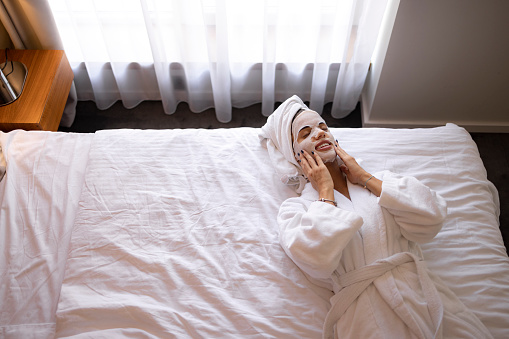 High angle view of young happy woman with facial mask relaxing during morning on a bed in bedroom.