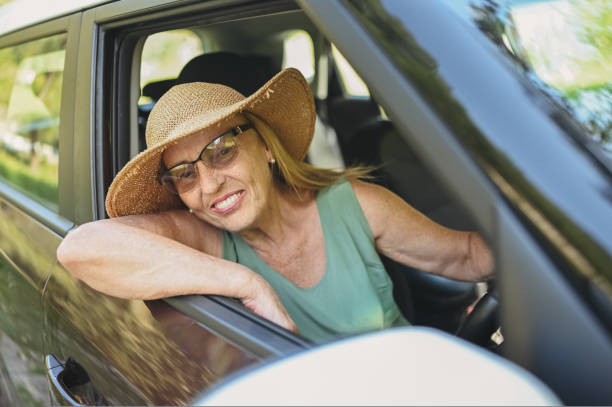 Happy senior woman driver in straw hat driving sitting in new car, smiling looking at camera enjoying journey. Driving courses and life insurance. Retired people activity and road trip concept. stock photo