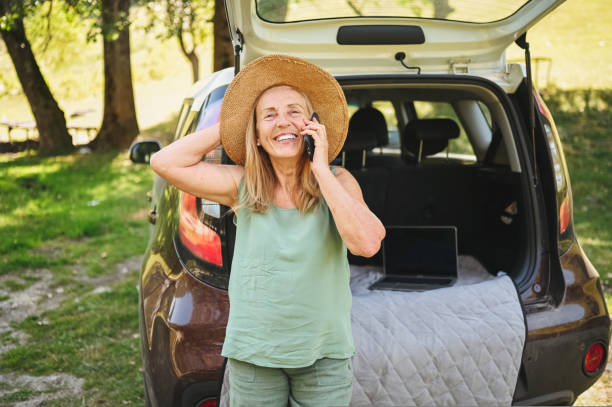 Senior active happy woman speaks phone car trunk or boot in camper sunny summer park - digital nomad freedom lifestyle concept, travel freelancer and vacations of retired old age people stock photo