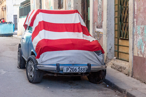 Havana, Cuba - October 27, 2022: A parked Polski Fiat car is covered with a large red and white striped cloth on the side of the street. A refuse bin, buildings and incidental people are in the background.