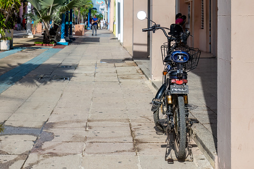 Holguin, Cuba - October 19, 2022: A bike parked by the side of a walkway next to an outside corridor with columns. Potted plants are on the other side of the walkway, and incidental people are on the scene.