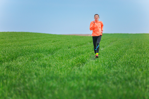 Young woman in orange windbreaker running on the  meadow on a clear day, cross country running