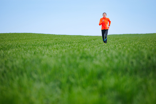 Young woman in orange windbreaker running on the green grass meadow on a clear day, cross country running