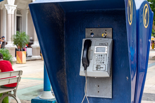 Holguin, Cuba - October 21, 2022: A coin-operated telephone in a phone booth in a park-like area. A building and incidental people are in the background.