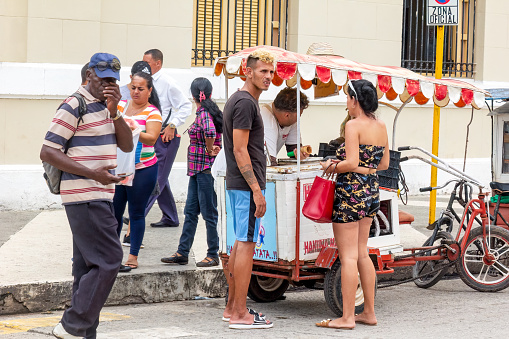 Holguin, Cuba - October 21, 2022: People standing around a bicycle street vendor. A building is in the background, and a signpost in front of the building says, 
