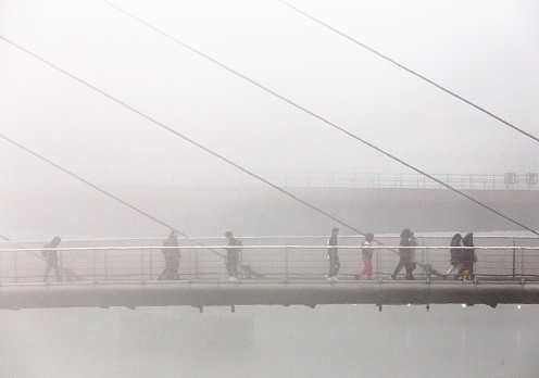 Monochrome image depicting commuters walking across a bridge on the Isle of Dogs in the docklands area of London, UK. It is a foggy day with low visibility, reducing the scene to an impressionistic one.