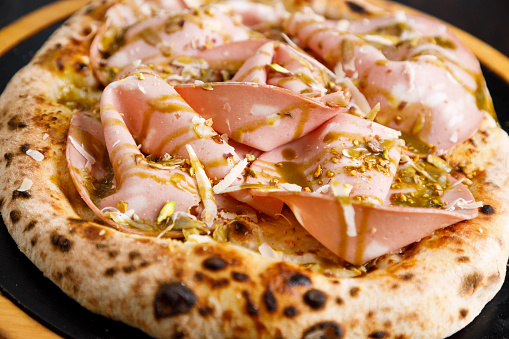 Close-up on baked pizza dough with fresh mortadella slices, mozzarella and pesto sauce condiment on top