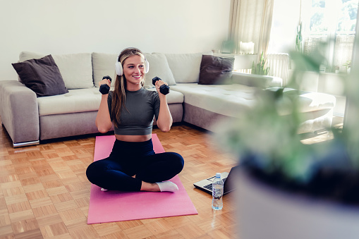 Shot of a sporty young woman using a laptop and doing lunges while dumbbells while exercising at home.