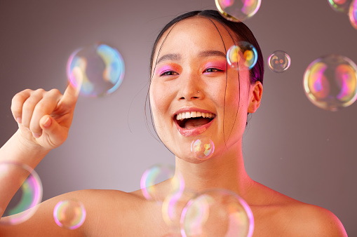 Beauty, bubbles and Asian woman with bright eyeshadow isolated on a studio background. Fun, glamour and colourful cosmetics with a beautiful Chinese woman popping bubbles for a healthy, fresh skin