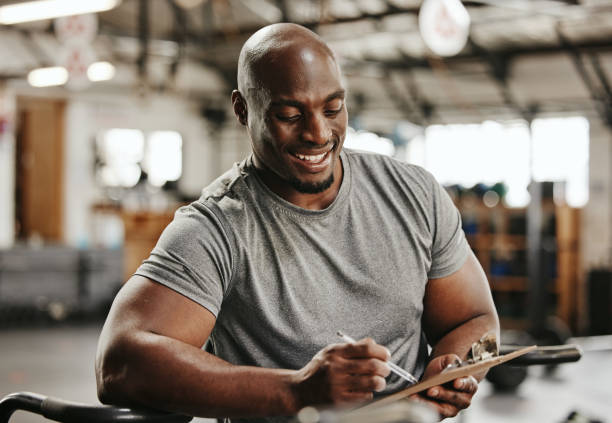 gym, workout and personal trainer with checklist on clipboard consulting for training sports in gym. black man of muscular, active and smiling fitness coach writing on health, wellness and exercise - men exercising equipment relaxation exercise imagens e fotografias de stock