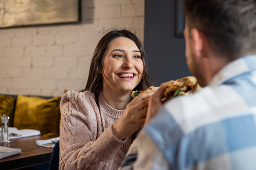 A young couple sitting at a wooden table in a modern restaurant, enjoying a meal out in Amble, North East England. They are both eating a chicken burger while looking at each other. The main focus is the woman looking at her boyfriend and smiling.