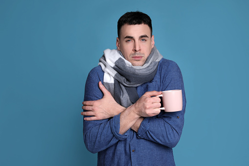 Young man with cup of hot drink suffering from fever on blue background. Cold symptoms