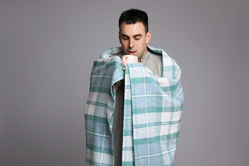 Young man wrapped in blanket with cup of hot drink suffering from cold on grey background