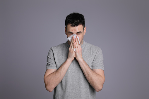 Young man blowing nose in tissue on grey background. Cold symptoms