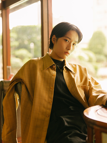 A half-body shot portrait of a young stylish or fashionable Asian woman with short hair wearing Autumn or Winter clothes, showing a cool attitude, sitting on  a chair around a table in front of the window with sunlight, in a western or vintage style room or cafe during daytime. She is looking at the camera for a photo.

A young Asian girl enjoying her day in a luxurious hotel indoors while relaxing during holiday or vacation.