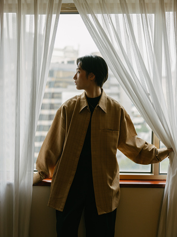 A half body shot portrait of a young stylish Asian woman with short hair wearing Autumn clothes, leaning against the window while looking at the view through the window between the curtains, in a room indoors during daytime. She is looking sideways at the view.

A young Asian girl enjoying her day in a luxurious hotel indoors while relaxing during holiday or vacation.