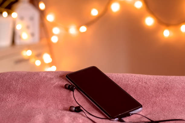 smartphone avec écouteurs connectés sur le bord du lit dans une ambiance cosy de lumières de noël. prêt à écouter la méditation. faible profondeur de champ. - at the edge of audio photos et images de collection