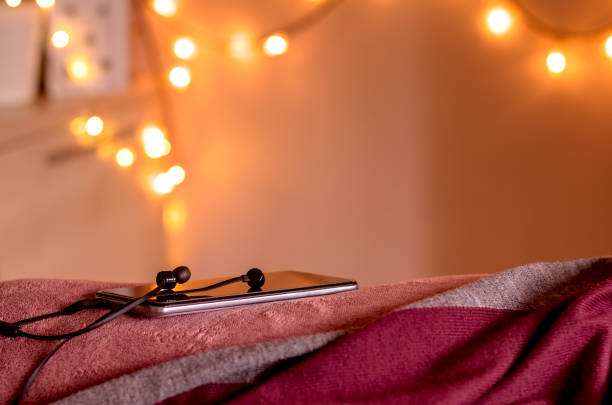 smartphone avec écouteurs connectés sur le bord du lit dans une ambiance cosy de lumières de noël. prêt à écouter la méditation. faible profondeur de champ. - at the edge of audio photos et images de collection