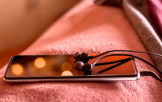 Smartphone with connected earphones on the edge of the bed in a cozy atmosphere of Christmas lights. Ready to listen to meditation. Shallow depth of field.