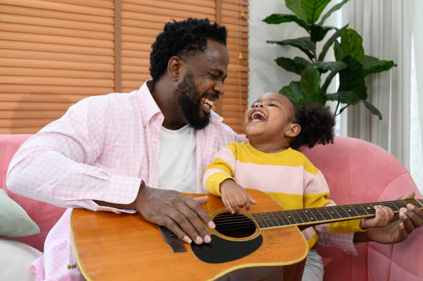 African girl and father are playing guitar while sitting on the sofa at home stock photo