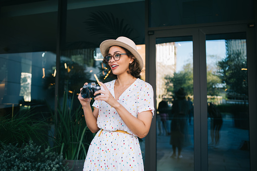 Positive woman in eyeglasses and trendy hat and casual white dress holding retro camera in hands while standing near glass door of modern building