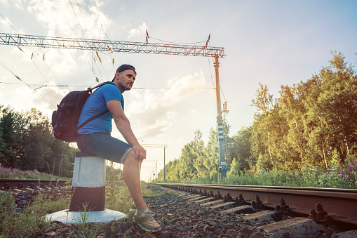 Tired male traveler with backpack seated on concrete pole on railtrack at sunset on summer time. Adventure and hiking concept.