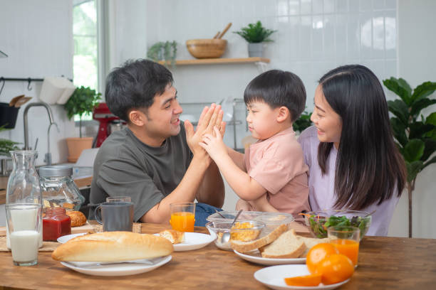 asian family breakfast at happy home. parents and children enjoy eating together, talking with laughter and good atmosphere. father plays with son playfully at kitchen table - domestic life young family family child imagens e fotografias de stock