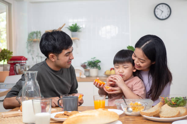 asian family breakfast at home on holiday. parents and children enjoy eating together, talking with laughter and good atmosphere. father plays with son playfully at kitchen table. - domestic life young family family child imagens e fotografias de stock