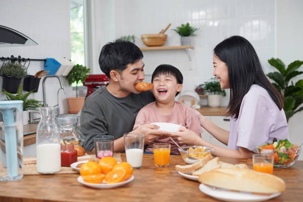 asian family breakfast at home. parents and children enjoy eating together, talking with laughter and good atmosphere. father and mother plays with son playfully at kitchen table. - two parent family indoors home interior domestic kitchen imagens e fotografias de stock