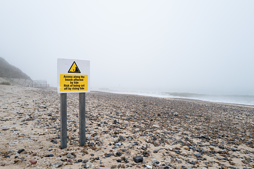 Yellow warning sign about a clear risk to life by being cut off by the rising tide of the North Sea. The beach is deserted due to being mid winter.