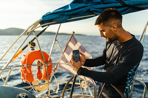 Young man at the helm while sailing on sailboat wearing a black sweatshirt and using mobile phone to take a photo of beautiful sunset
