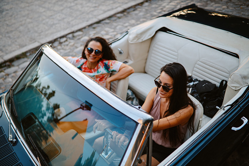 Female friends going out for a joyride in the evening, having a fun weekend ahead. They’re driving in a convertible vintage car, having time for themselves.