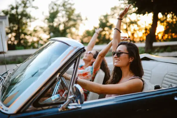 Female friends going out for a joyride in the evening, having a fun weekend ahead. They’re driving in a convertible vintage car, having time for themselves.