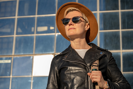 30s woman with short blonde hair in leather jacket and trendy hat standing next to the modern building in Barcelona